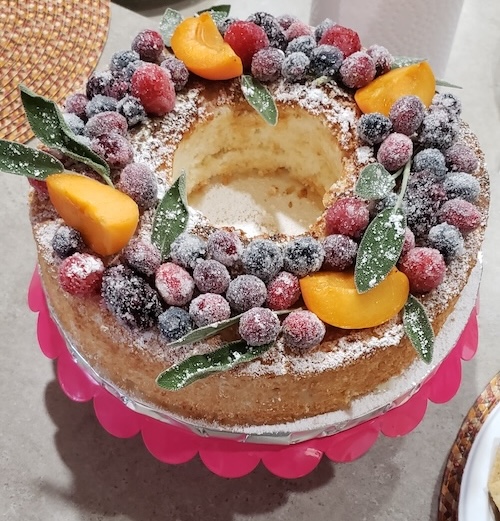 Image of a round bundt-styled vanilla cake powdered with confectioner sugar, decorated with sugared berries with sliced fruit and herbs.