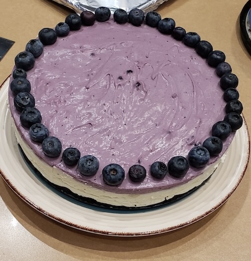 Image of purple blueberry cheesecake on a plate, decorated with blueberries in a circle around the edges of the cheesecake.